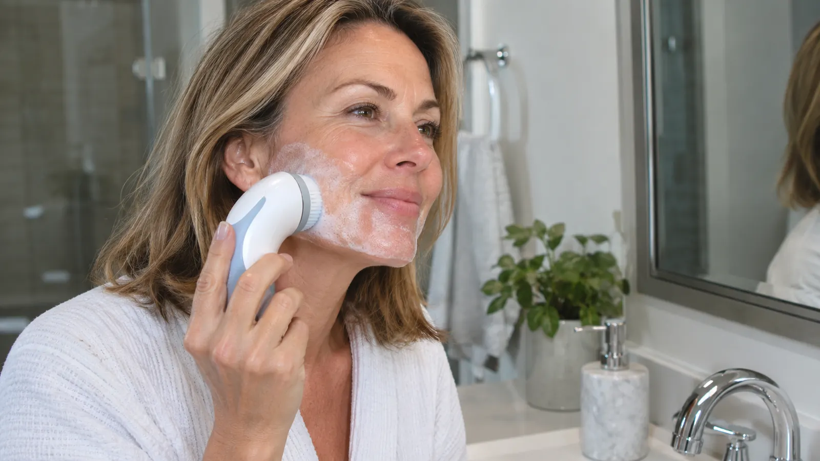 A woman in her late 40s using a facial cleansing brush on her cheek with cleanser foam on her skin