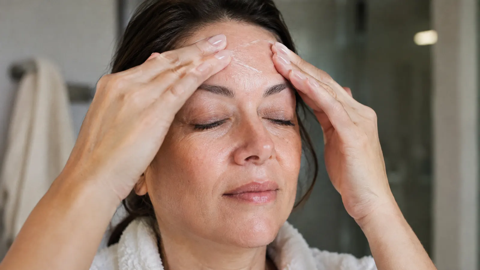 A woman in her late 40s gently pressing moisturizer onto her forehead with her ring fingers, eyes closed