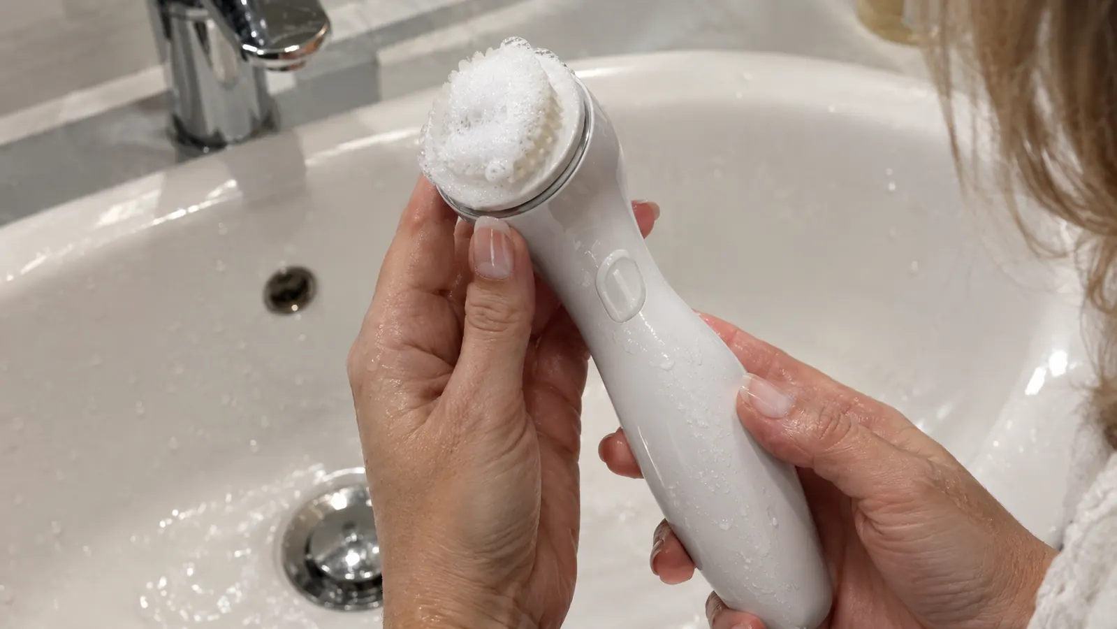 A hand holding a facial cleansing brush with foamy cleanser on the bristles over a sink