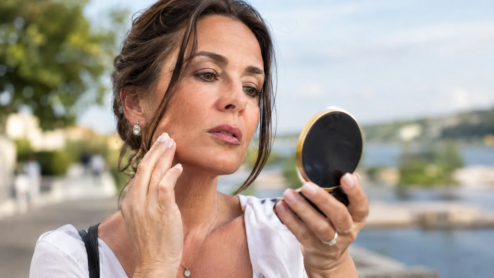 A woman in her late 40s checking her face in a compact mirror outdoors in daylight