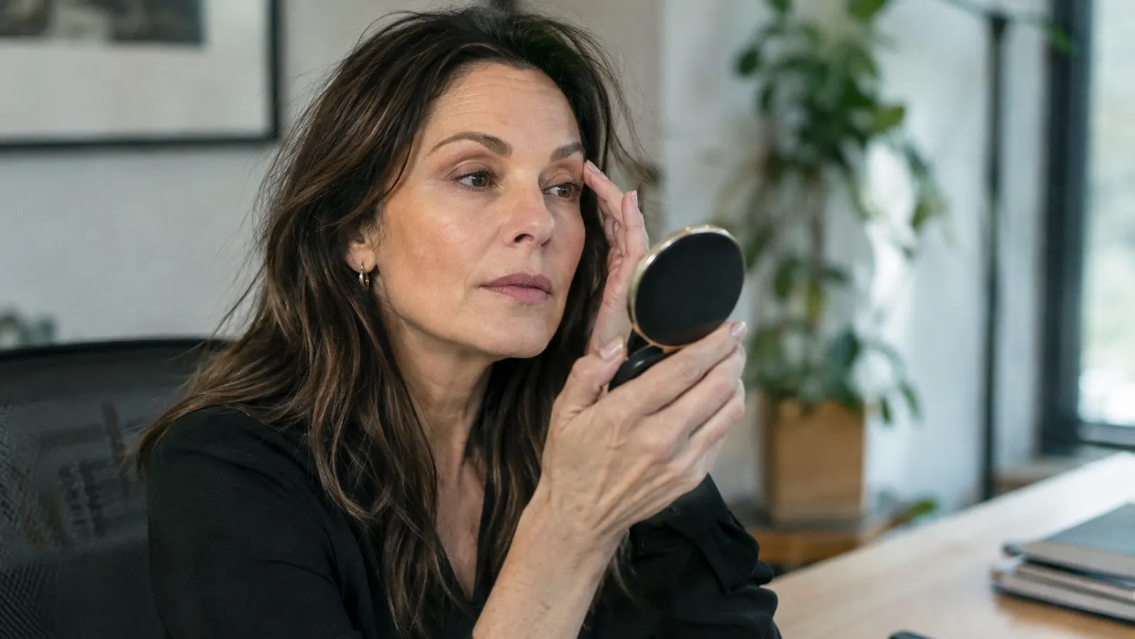 A woman in her late 40s checking her lashes in a compact mirror at a desk in the afternoon