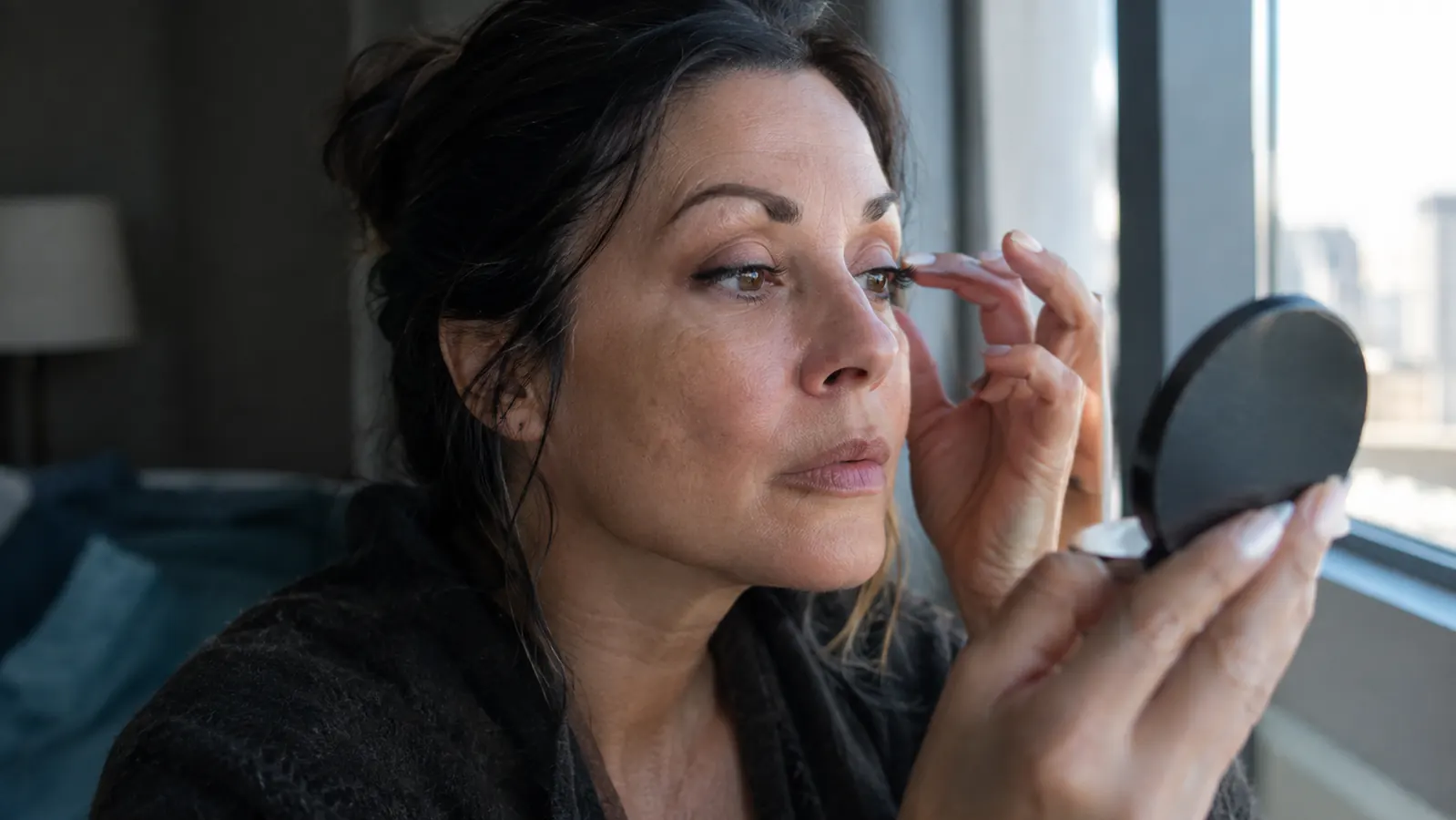 A woman in her late 40s looking at her reflection in a compact mirror, natural daylight