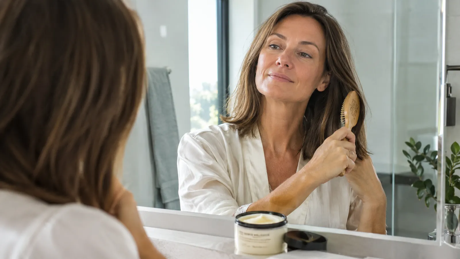 A woman in her late 40s brushing dry, smooth shoulder-length hair in front of a bathroom mirror, a jar of hair mask on the counter